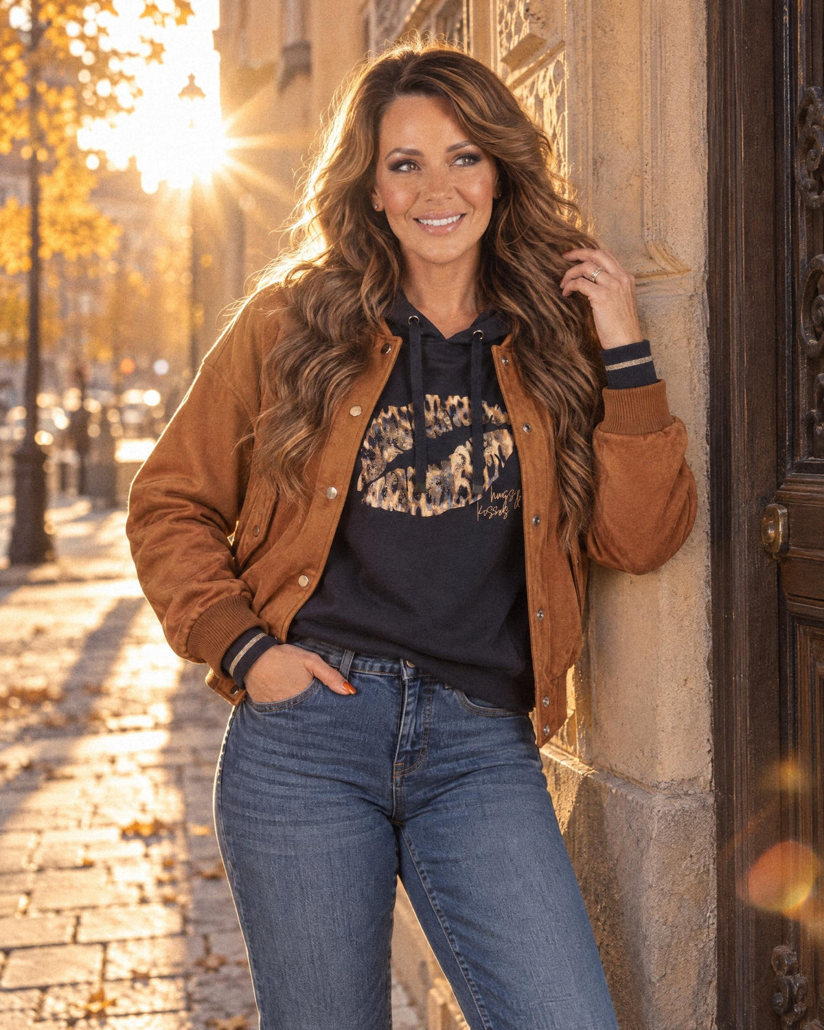 Sofie i brun mockajacka från Betty Barclay, mörkblå tröja med läppmotiv från Betty Barclay och medelblå jeans från Cambio mot en solig husfasad