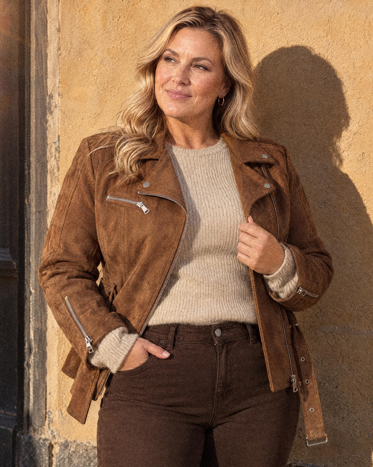 Milla i brun mockajacka från Missy, sandfärgad tröja från Haust och bruna jeans från Frank Lyman mot en solig vägg